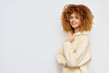 Yumuşak krem süveter giyen, neşeli, kıvırcık saçlı genç bir bayan, zarif bir şekilde arka planda duruyor, pozitif enerji yayıyor ve arkadaş canlısı bir hava veriyor.