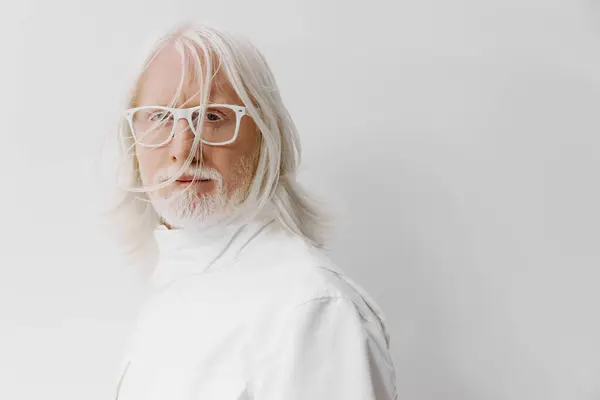 stock image An elderly man with bright white hair and stylish glasses poses against a soft, neutral backdrop His outfit is all white, reflecting a contemporary aesthetic and a blend of classic and modern trends