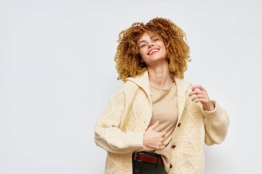 Kıvırcık saçlı neşeli bir genç kadın, rahat krem renkli bir kazakla bir anın tadını çıkarıyor. Yumuşak ışık arka planı, hayat tarzı ve moda görselleri için ideal olan neşeli ifadesini geliştiriyor.