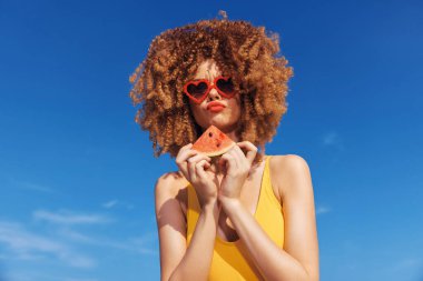 Sarı mayo giymiş neşeli bir kadın karpuz dilimiyle poz veriyor, kalp şeklinde güneş gözlüğü takıyor, yaz havası yayıyor.
