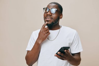 Gözlüklü, beyaz tişörtlü, düşünceli bir genç adam akıllı bir telefon tutuyor. Yüzünde hafif bir gülümseme var. Yumuşak ve tarafsız bir arkaplan oluşturuyor.