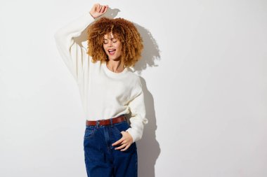 Kıvırcık saçlı, beyaz kazaklı, mavi kotlu, açık arkaplanda dans eden neşeli bir kadın. Bu görüntü hayat tarzı teması için mükemmel, coşkulu ve tasasız bir ruh hali yakalar.