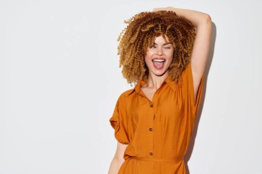 Modern turuncu kıyafetli neşeli kıvırcık saçlı kadın geniş bir gülümseme sergiliyor, temiz ve nötr bir arkaplanda yakalanmış görüntü eğlence, mutluluk ve moda simgesi.