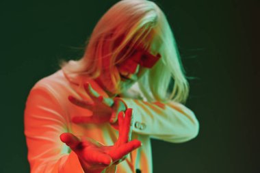 Kırmızı gözlüklü, uzun beyaz saçlı şık bir şahıs, sanatsal ifadeyi vurgulayan çağdaş bir estetiği somutlaştıran canlı yeşil bir arka plana çarpıcı bir şekilde karşı durmaktadır.