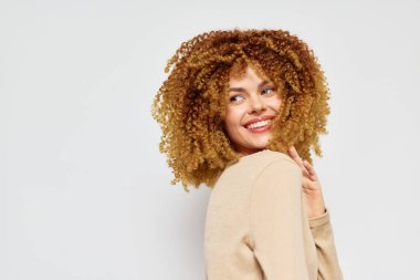 Şehvetli, kıvırcık saçlı gülümseyen genç kadın açık gri bir arka planda poz veriyor. Görüntü neşe, kendine güven ve doğal saç stillerinin güzelliğini yansıtıyor. Yaşam tarzı için mükemmel.