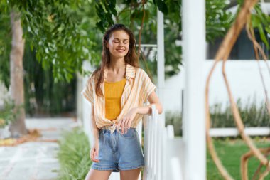 Bereketli bir bahçede beyaz çitin yanında duran neşeli genç bir kadın açık sarı bir bluz giyiyor, yeşil yapraklarla çevrili, canlı, kaygısız bir hava yayıyor.