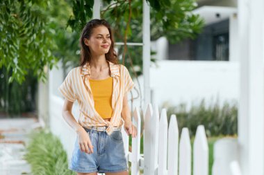 Beyaz bir bahçe çitinin yanında neşeli bir yüzü olan, sarı bluz ve kot şortla bembeyaz çitlerle çevrili genç bir kadının olduğu sıradan moda sahnesi görsel olarak çarpıcı.
