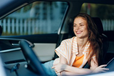 Uzun saçlı neşeli genç bir kadın rahat bir şekilde arabada oturuyor, rahat bir kıyafet giyiyor. Parlak bir gülüşü var, mutluluk ve rahatlık taşıyor. Arka planda hafif bulanık bir açık hava var.