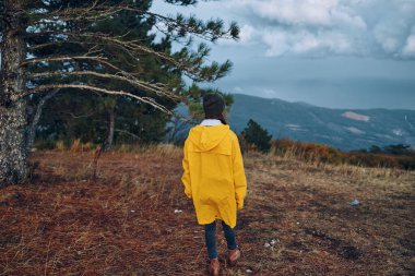 Sarı yağmurluklu bir kadın arka planda görkemli dağlarla pitoresk bir tarlada duruyor, seyahat ve açık hava macerası güzelliğinin vücut bulmuş hali.