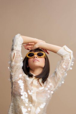 Sarı güneş gözlüklü Asyalı bir kadın ve bej arka planda beyaz bir bluz. Elleri başının üstünde, rahat ve şık bir havayı somutlaştırıyor.