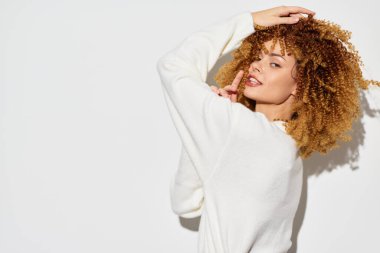 Şık, genç bir kadın, rahat beyaz bir kazak giyerken kıvırcık saçlarını sergiliyor. Minimalist beyaz bir arkaplana karşı neşeli bir şekilde poz veriyor. Kendine güveni ve çekiciliği var.