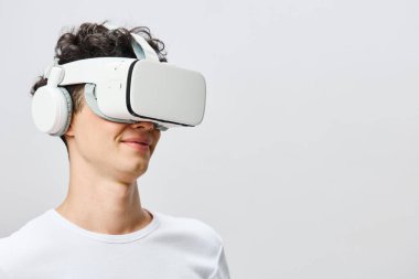 Genç bir adam sanal gerçeklik deneyimine dalmışken gülümseyen bir VR kulaklık ve kulaklık takıyor. Beyaz arka plan fütüristik teknolojiyi ve kullanıcıların katılımını vurguluyor.