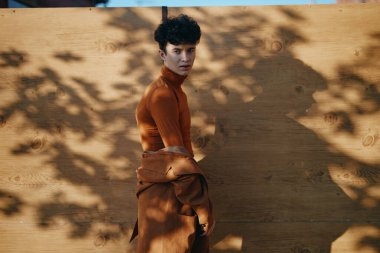 Modayı takip eden bir birey, kahverengi bir kıyafetin içinde, ahşap arka planın sıcak tonlarını yükselterek sanatsal bir gölge etkisi yaratıyor.