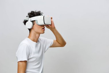 Genç adam sanal gerçeklik deneyimi yaşadı, beyaz bir VR kulaklık ve kulaklık takıyor, minimal gri bir arka plan ile kurulmuş, modern teknoloji ve büyüleyici oyun kavramları sergiliyor.