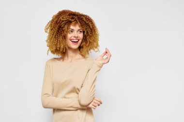 Şehvetli kıvırcık saçlı, bej bir kazak giyen, yumuşak bir arka planda duran mutlu genç kadın. Neşeli ifadesi güven ve pozitifliği yansıtıyor. Güzellik ve moda için mükemmel.