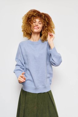 Moda, manken, neşeli, mavi süveter, yeşil etek, günlük tarz, kıvırcık saçlı mutlu bir kadın mavi kazak ve yeşil etek giyer, minimalist beyaz bir arkaplan önünde pozitiftir.