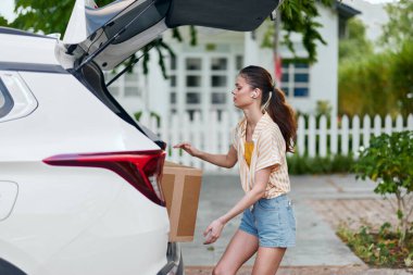 Gündelik giysiler içindeki genç bir kadın beyaz bir arabanın bagajından karton bir kutu boşaltıyor dekor parlak ve açık, yeşil yapraklı ve yerleşim alanı olan, canlı bir arka plan sergiliyor.