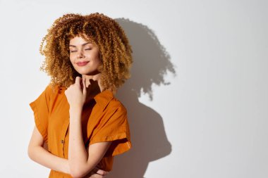 Turuncu giysili kıvırcık saçlı genç bir kadın, sade bir arka planda şakacı pozlar veriyor. Kendine güveni ve yumuşak gölgesi görüntüye derinlik katıyor.
