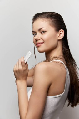 Pürüzsüz, ıslak saçlı bir kadın elinde tarak tutarken gülümsüyor, minimalist bir ışık arkaplanının karşısında bu görüntü güzellik, saç bakımı ve kendine güvenin kavramlarını vurguluyor.