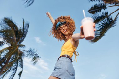 Sarı bluzlu ve kot etekli neşeli bir kadın smoothie tutuyor, güneşin altında, canlı palmiyeler ve açık mavi gökyüzü tropikal havayı güçlendiriyor.
