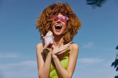 Güneş gözlüklü neşeli bir kadın dondurma tutuyor, açık mavi bir arka plana gülümsüyor.