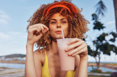 Dalgalı saçlı ve renkli vizörlü bir kadının okyanus kenarında ferahlatıcı bir içkinin tadını çıkardığı tasasız bir sahne.