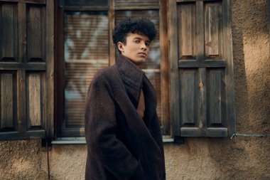 Kıvırcık saçlı, kahverengi paltolu moda adam kepenkli pencereli ve ahşap kapılı eski binanın önünde duruyor..