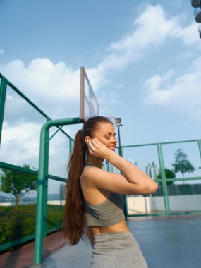 Gülümseyen fitness meraklısı uzun saçlı bir basketbol sahasında çalışırken mavi gökyüzü arka planıyla müzik dinliyor.