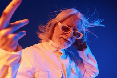 Uzun beyaz saçlı ve güneş gözlüklü havalı yaşlı bir adam çarpıcı bir mavi arka plana karşı poz veriyor. Oyuncu tavrı ve eşsiz stili sanatsal açıdan mükemmel ve çekici bir atmosfer yaratıyor.