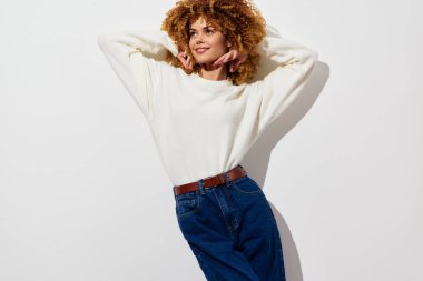 Sade beyaz bir kazak ve yüksek bel kot pantolon giyen şık genç bir kadın model, minimalist beyaz bir arka plana karşı poz veriyor, kendine güveni yüksek ve modaya uygun bir hava yayıyor.