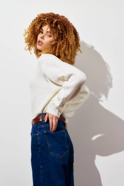 Sarışın, kıvırcık saçlı, minimalist beyaz arka planda poz veren, sıcak beyaz bir kazak ve mavi kot giyen şık ve kendine güvenen bir kadın.