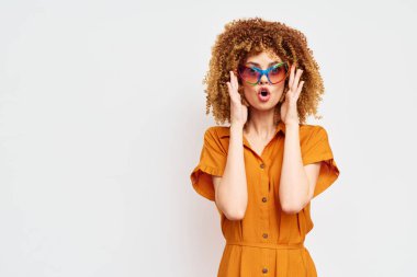 Kıvırcık saçlı, parlak güneş gözlüklü, modaya ve kişisel tarzına dikkat çeken, minimalist açık bir arkaplan karşısında şaşırmış bir ifade takınan modaya uygun genç bir kadın.