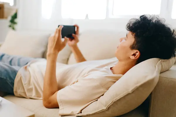 stock image young man comfortably lying on a sofa, using a smartphone in a bright living room setting, showcasing a relaxed lifestyle, modern decor, and the enjoyment of leisure activities with natural light