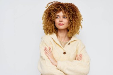 Şehvetli kıvırcık saçlı ve neşeli yüzlü, açık bej bir kazak giymiş, minimalist beyaz bir arka plana karşı poz vermiş, pozitiflik ve rahatlık saçan kendine güvenen genç bir kadın.