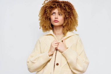 Zarif genç bir kadın, yumuşak krem rengi hırka giyerken güzel kıvırcık saçlarını sergiliyor. Görüntü, ifadesine ve tarzına odaklanan sade bir arkaplan içeriyor.