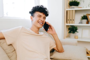 Kıvırcık saçlı neşeli genç bir adam, parlak ve havadar bir oturma odasındaki koltukta rahat bir şekilde otururken telefon konuşmasından zevk alıyor. Modern dekorda yeşil bitkiler ve organize raflar yer alıyor.