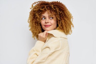 Şehvetli kıvırcık saçlı, şık, rahat bir süveter giyen, omzunun üzerinden rahatlık ve cazibe saçan bir arka plana bakan neşeli bir genç kadın.