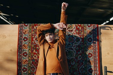 Genç bir adam, modern moda estetiğini vurgulayan modaya uygun giyinmiş, güzel desenli bir halının önünde dururken kollarını uzatıyor.