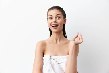 Havluya sarılmış, mutlu, genç bir kadın, beyaz bir arka plana sarılmış, heyecan ve pozitiflik yayıyor. Hareketleri ve ifadesi görüntüye neşe ve neşe katıyor.