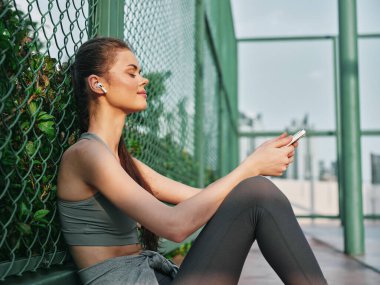 Aktif giyimli genç bir kadın telefonuyla müzik dinlemekten hoşlanır. Açık havada yeşil bir çite yaslanır.