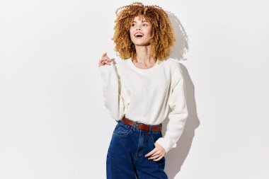 Şık, kıvırcık saçlı, rahat beyaz bir kazak ve yüksek bel kot pantolon giyen, gülümseyen ve hafifçe poz veren genç bir kadın. Temiz bir görünüm için.