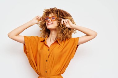 Kıvırcık saçlı şık bir genç kadın turuncu bir elbise ve temiz beyaz bir arka plana göre ayarlanmış gözlük takarak neşeli bir poz veriyor, kendine güveni modern yaz modasını simgeliyor.