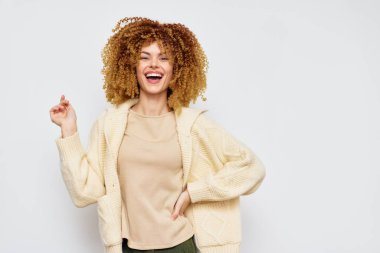 Dolgun kıvırcık saçlı neşeli bir kadın, rahat bir fildişi süveter giymiş, geniş bir tebessüm ile hafif bir arka plana karşı gülümsüyor yaşam tarzı ve çeşitli moda kavramları için ideal.