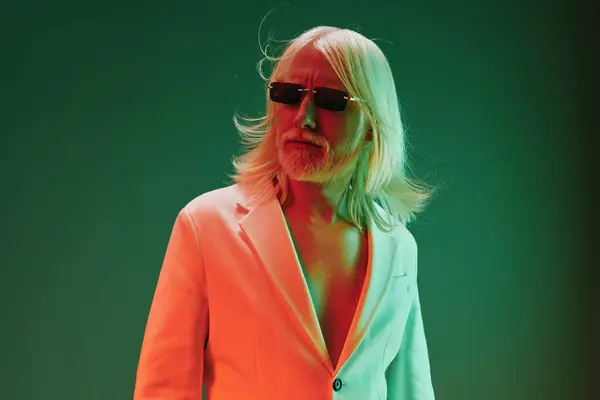 stock image A fashionable older man with long white hair and sunglasses stands confidently in front of a glowing green and orange backdrop, embodying a modern aesthetic His stylish white suit enhances the