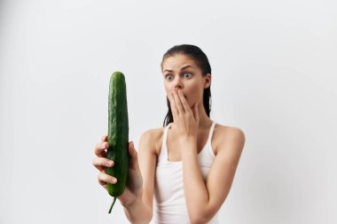 Beyaz atletli genç bir kadın yüzüne salatalık tutarken şaşkına döner. Tarafsız arka plan onun neşeli ifadesini güçlendirir ve neşeli bir atmosfer yaratır.