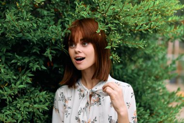 Canlı, kızıl saçlı bir kadın yemyeşil çalıların önünde dikiliyor.