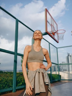 Neşeli bir kadın bir basketbol sahasında duruyor, spor sutyen ve bağlı bir gömlek giyiyor, parlak bir gökyüzüne karşı.