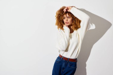 Moda mankeni, beyaz bir süveter ve mavi kot pantolon içindeki kıvırcık saçlarıyla temiz beyaz bir arka plana karşı durarak tarz ve çağdaş moda eğilimleri sergiliyor.