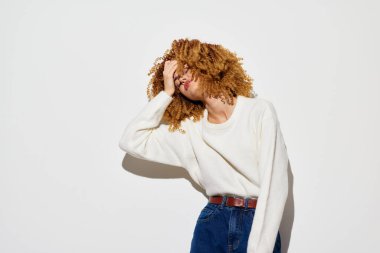 Kıvırcık saçlı neşeli bir kadın, rahat beyaz bir kazak ve kot pantolon giymiş, elini şakacı bir şekilde alnına koyuyor, temiz beyaz bir arkaplanda yakalanmış.