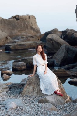 Beyaz elbiseli sakin kadın kayalık sahilde oturuyor. Arka planda su ve kayalar var.